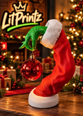 3D-printed Grinch arm ornament holder dangling a red ornament beside a Christmas tree
